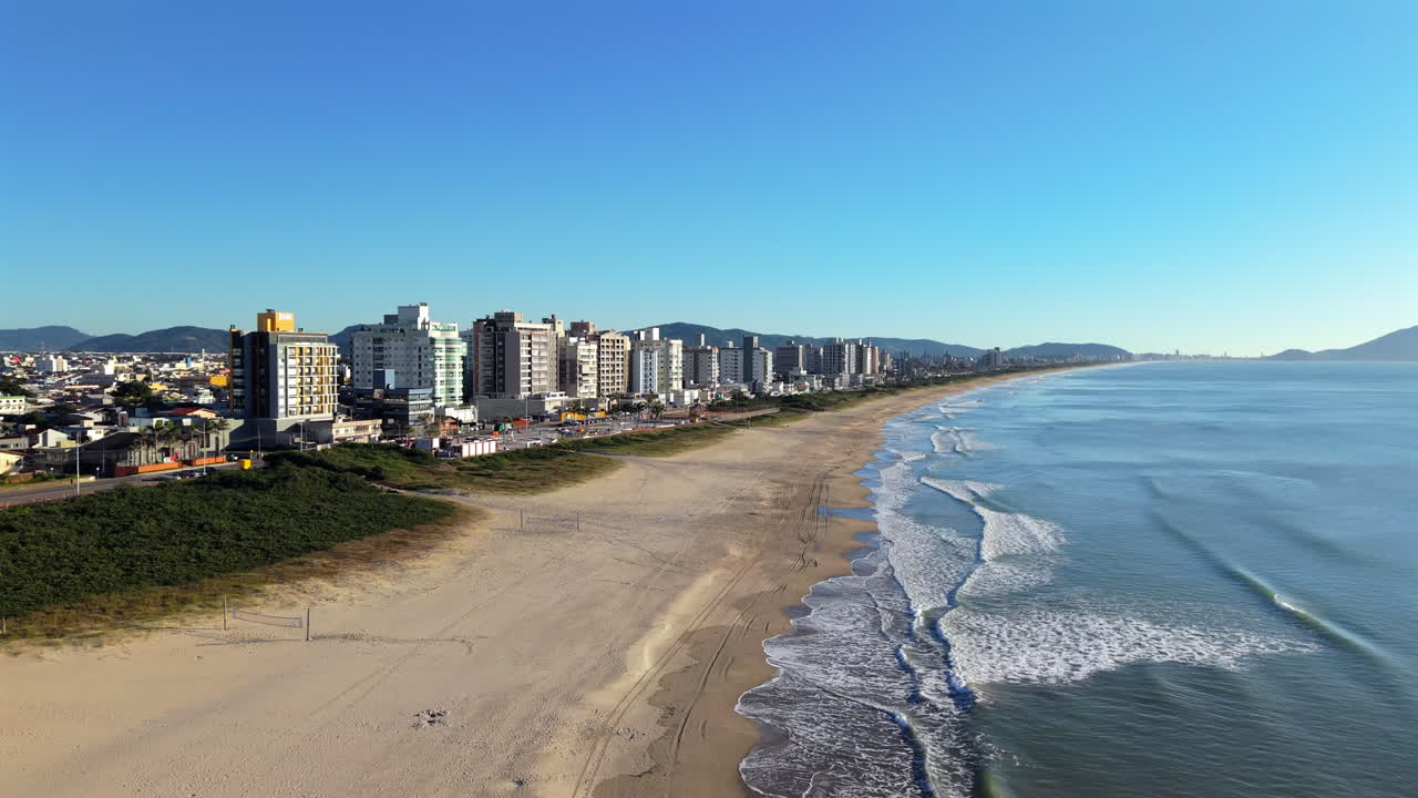 Navegantes beach in Santa Catarina, Brazil, showcasing hotels, golden sand, and the rhythmic crashing of ocean waves on a sunny day, stunning aerial glide