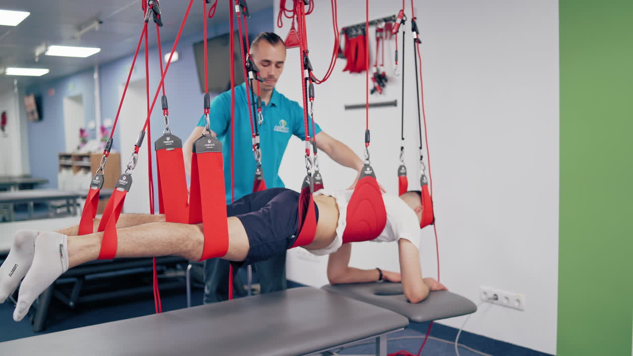 Man's body is hanging on the special suspension ropes on the modern rehab center background. Personal trainer helps the man to do the exercises with the new equipment to recover.