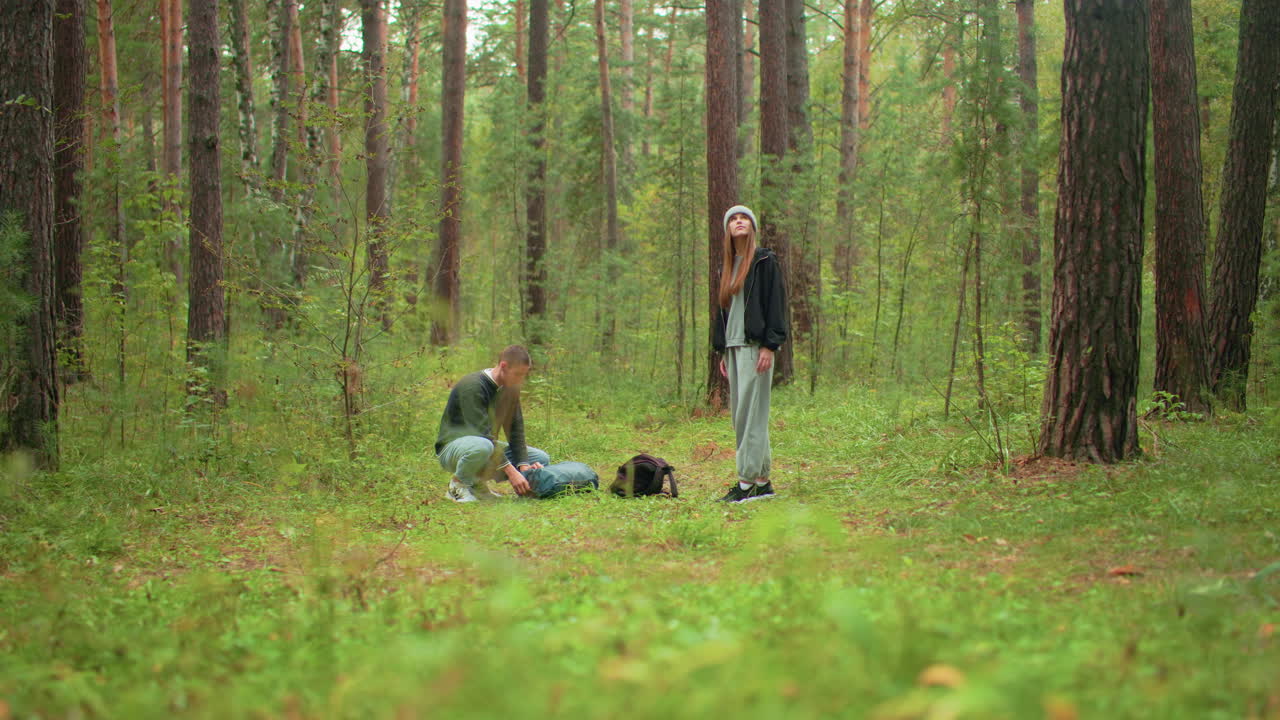 campers in woodland as man crouches to open green travel bag while woman stands looking around thoughtfully among tall trees and green foliage, preparing to set up temporary camp in forest area