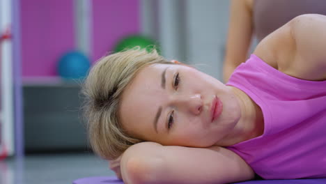 close-up of lady in pink working out on mat with gym trainer assisting, focused on stretching, with exercise ball visible in modern gym background