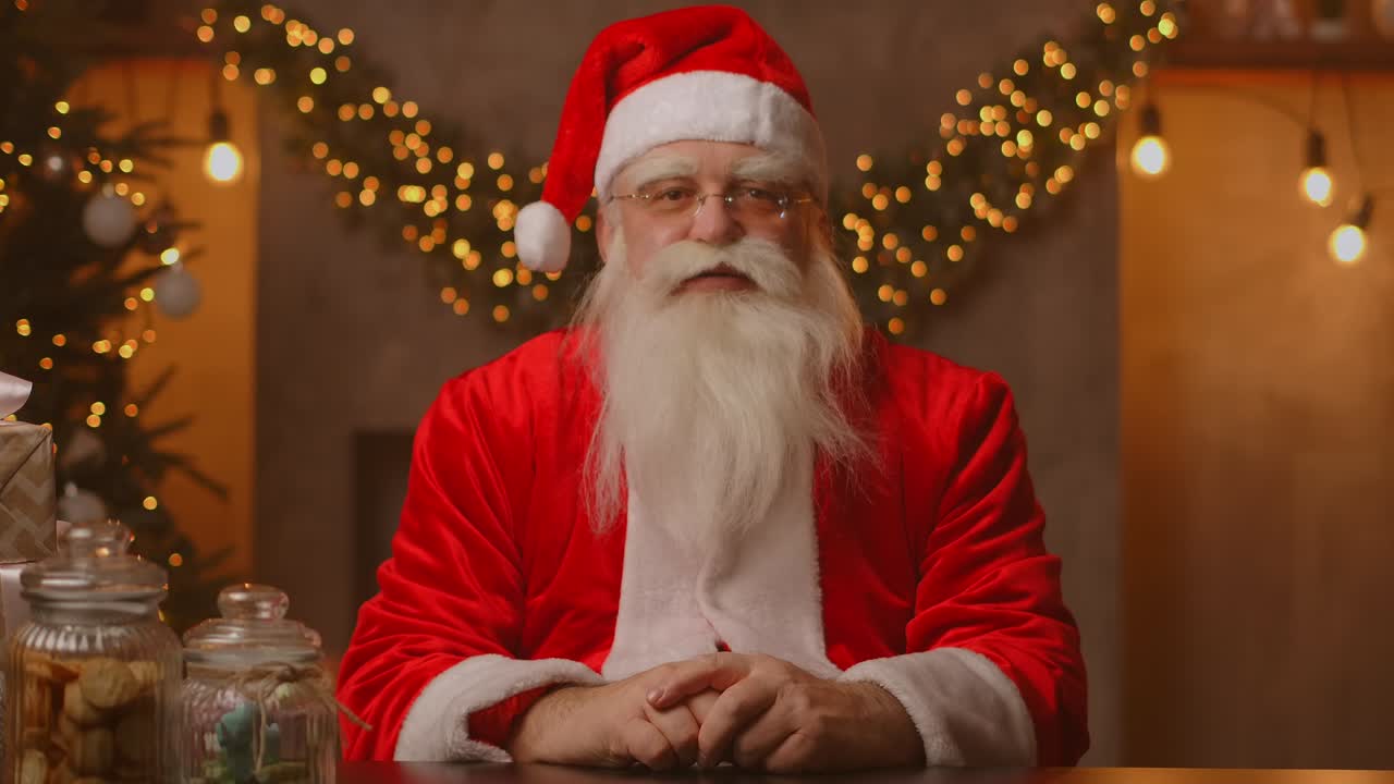 산타클로스는 크리스마스를 축하하기 위해 장식된 아름다운 거실의 테이블에 앉아 있으며 오른손을 흔들고 말을 시작합니다.