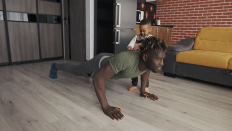 dad in casual clothes doing push-ups at home, little son helps him holding shirt