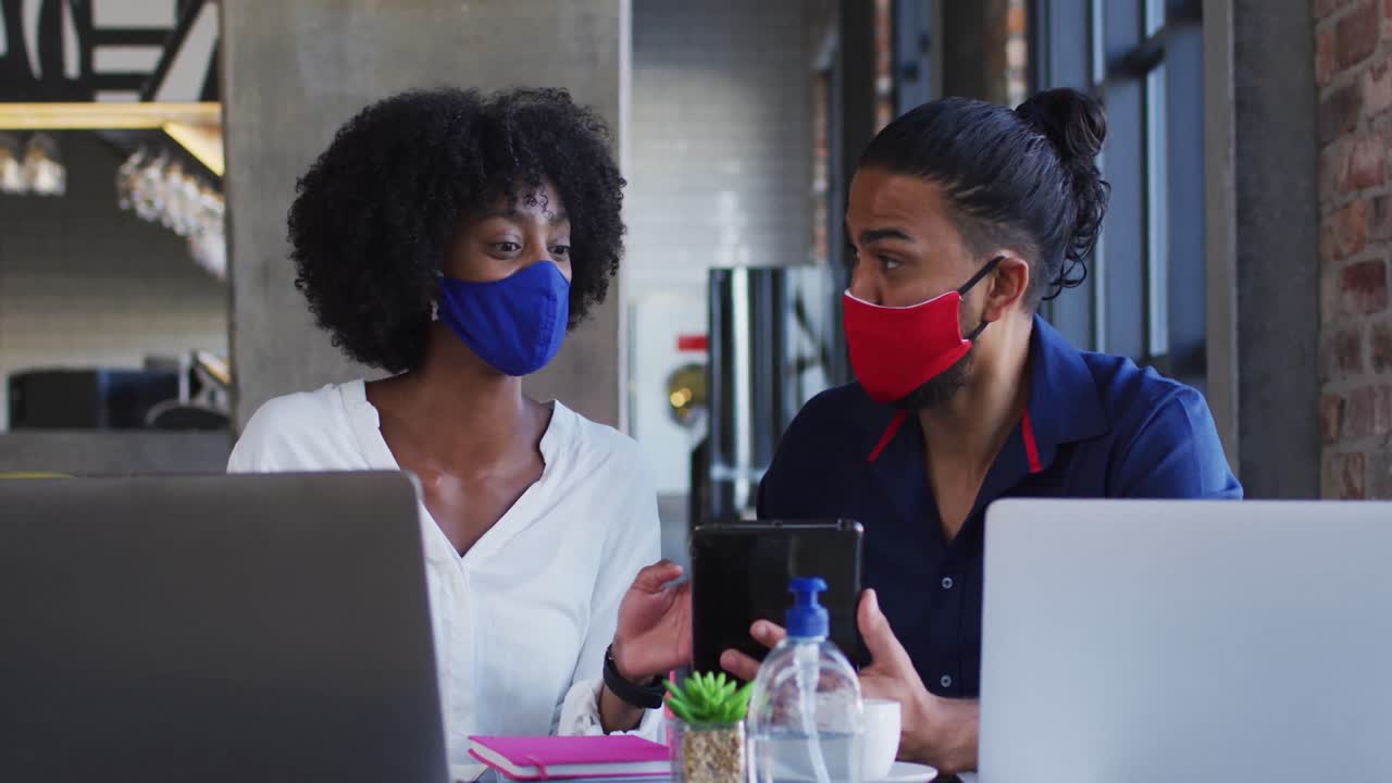 Diverse couple wearing face masks sitting in cafe using laptops and smartphone