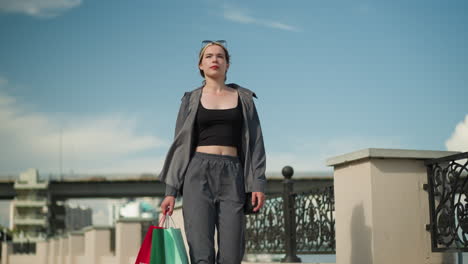 white lady in casual grey outfit walking near a bridge fence, holding three colorful shopping bags, glancing to the right. background shows cars passing on the overhead bridge with clear blue sky