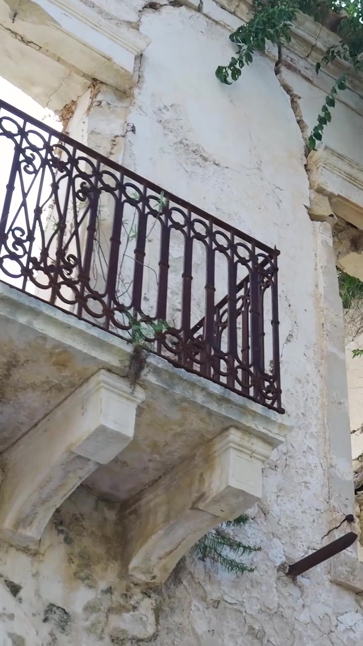 Video verticale che inquadra una parete bianca assolata e scrostata di un edificio abbandonato a Xylokastro, con intonaco crepato, ringhiera del balcone in ferro e vegetazione incolta. Il degrado urbano incontra il fascino mediterraneo