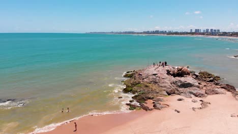 drone flying left to right showing some people fishing on the rock in the shore. some other fisherman walks in the sand