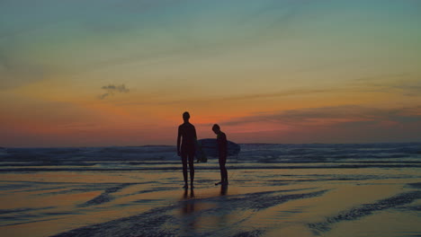 two surfer silhouettes on vibrant beach during rainbow sunset