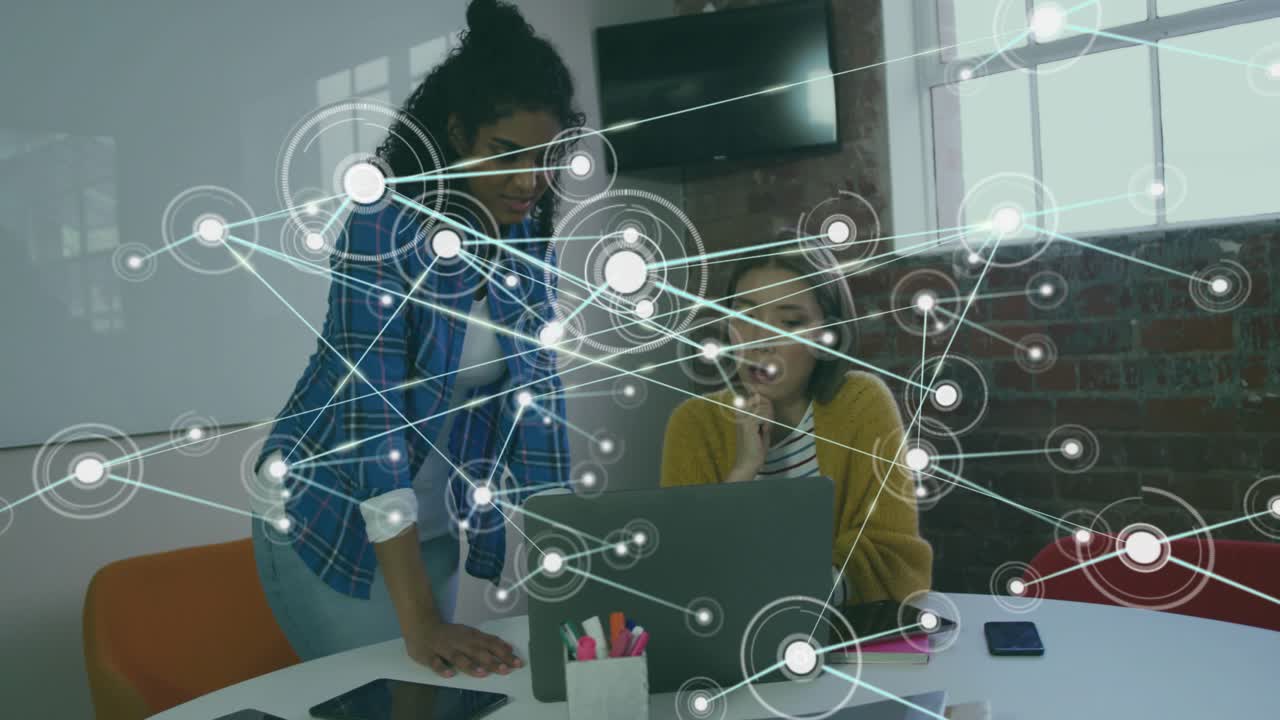 Two women leaning at laptop in business meeting pointing and causing network overlay showing data