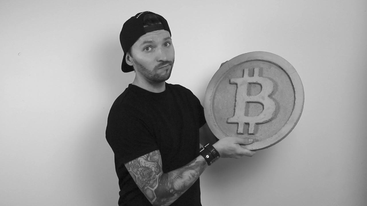 Gråtoneoptagelse af en tatoveret mand, der løfter en stor beton- eller sten-bitcoin-skulptur og ryster på hovedet i misbilligelse, mens han er utilfreds. Kryptovaluta løftes i slowmotion af manden.