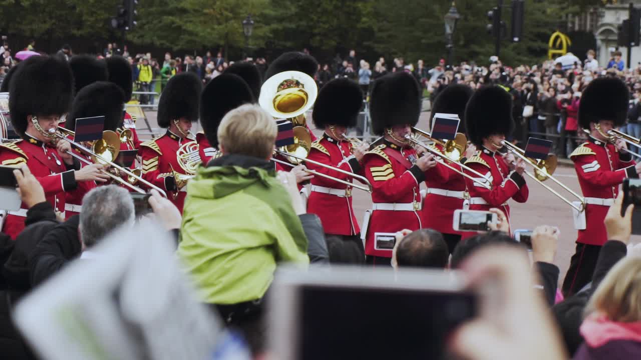 London Queen's Guard black-hatted redcoats play musical instruments and march in procession with spectators watching and taking photos, handheld close up slow motion