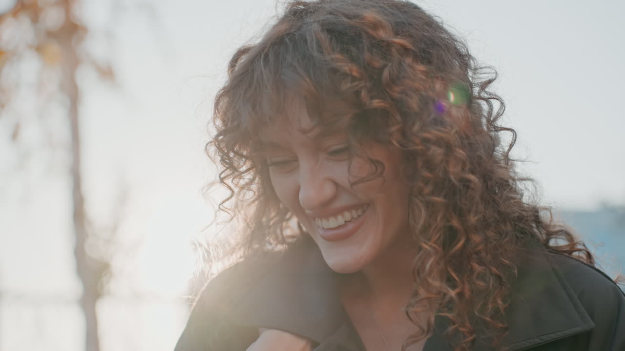 Sguardo dolce di profilo di donna con capelli ricci e cappotto vicino a una recinzione di ferro. Il controluce del tramonto, lo sfondo cittadino attenuato e un delicato effetto bokeh creano un'atmosfera sognante e introspettiva. Sorriso sottile e postura composta.
