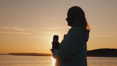 Lonely-Woman-Drinks-Beer-From-A-Can-On-The-Lake-Standing-Alone-By-The-Fence-Looking-At-The-Sunset