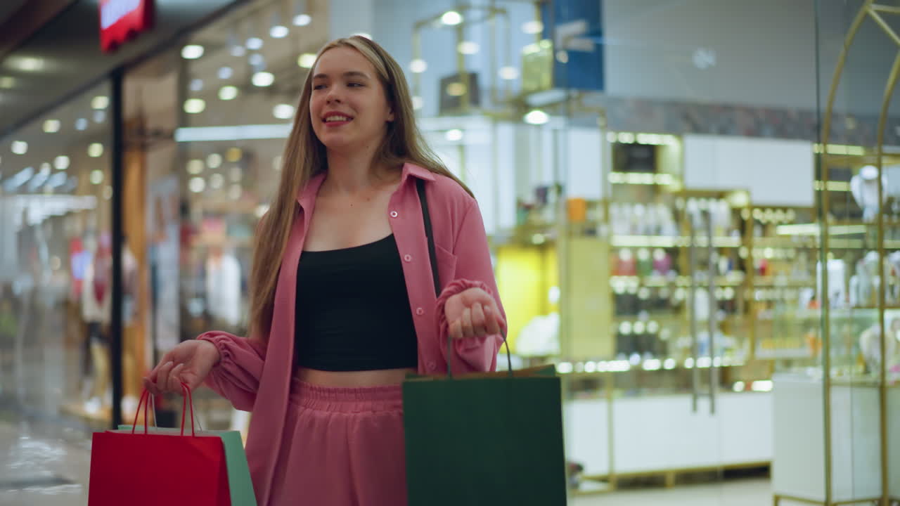 dama de vestido rosa y pantalones cortos sosteniendo bolsas de compras rojas, verdes y verdes con una sonrisa brillante en un centro comercial, el fondo está ligeramente borroso con una vista suave de compradores y tiendas