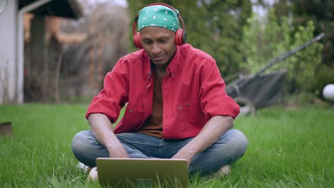 retrato frontal de un joven guapo feliz y sonriente disfrutando de la música en los auriculares mirando la pantalla de la computadora portátil sentado en la hierba verde en el jardín. florista relajado descansando al aire libre. jardinería y descanso.