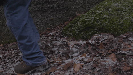 view of feet only of a man wearing hiking boots and blue jeans jumps from a mossy rock onto the leaf covered forest floor