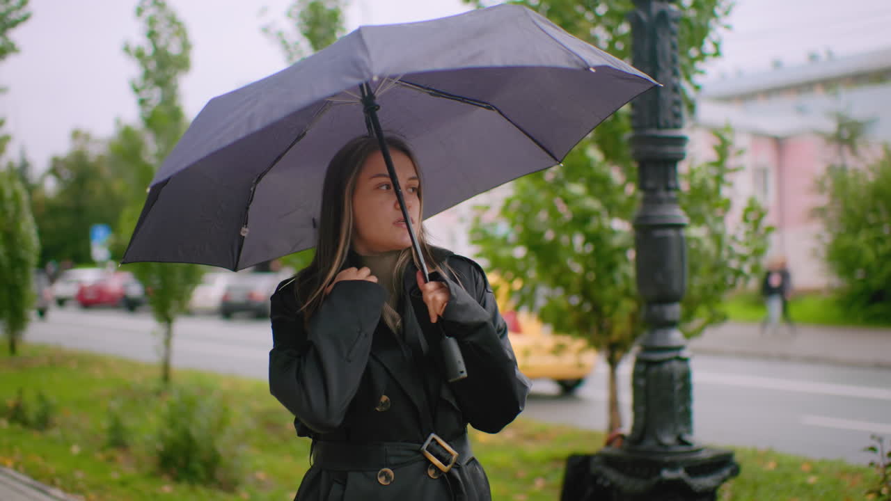Pretty girl in black trench coat holding umbrella standing on city street sidewalk during cold overcast day with background of greenery cars and buildings showing calm thoughtful outdoor lifestyle