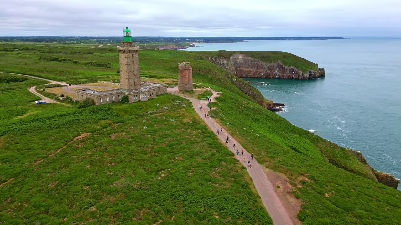 Tourists walking path towards iconic Cap Fréhel lighthouse and old tower on green headland, Brittany, France. Aerial forward