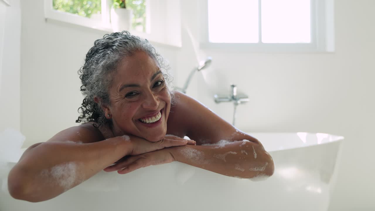 Senior woman leaning on tub rim, suds, camera zooming, smiling and laughing at lens in bathroom