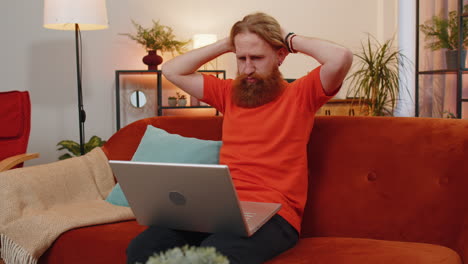 bearded redhead man use laptop surprised by bad news, fortune loss, fail, lottery results at home