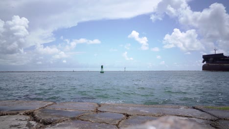 An-industrial-ship-floats-into-the-channel-between-jetties-as-waves-lap-up-onto-the-granite-rocks-in-Port-Aransas,-Texas-on-the-Gulf-of-Mexico