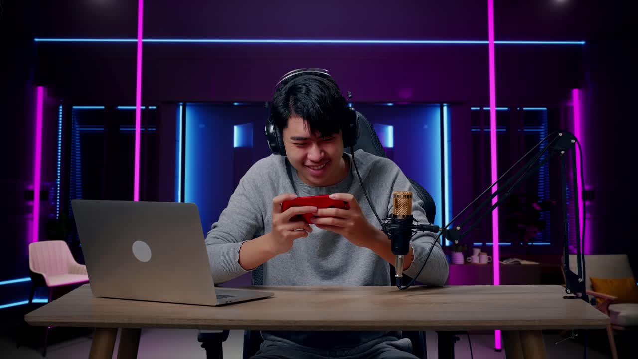 un hombre asiático sonriente con auriculares y computadora usando un teléfono móvil jugando un juego en una habitación iluminada por luces de neón en retro