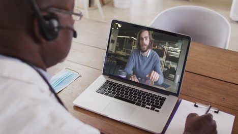 african american male doctor wearing phone headset taking notes while having a video call on laptop