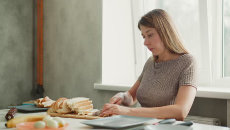 medium shot of griller slicing bread into bite sized pieces on wooden board, table arranged with sausage banana eggs bowl knife under bright window light capturing calm cooking preparation scene