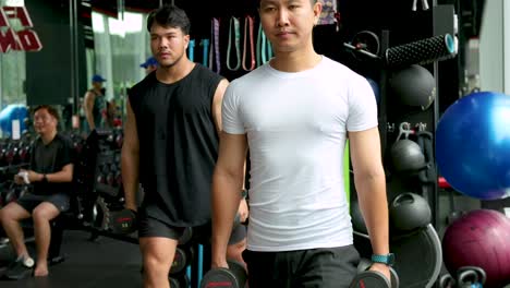Two men exercise with dumbbells in a well-equipped gym, showcasing determination and strength under bright lighting