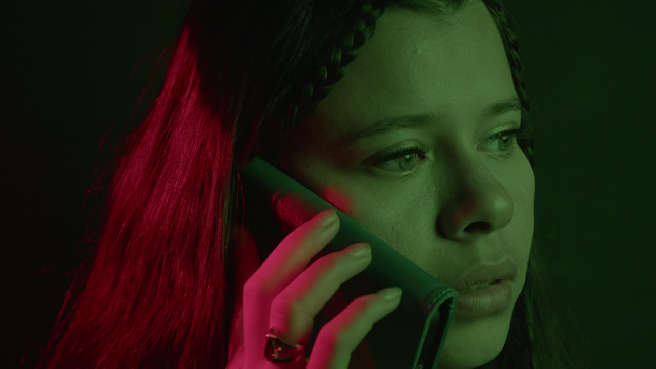 Emotive close-up of a woman on a phone call, illuminated by moody green and red lighting, conveying tension and intrigue. The shot captures intense focus and expression