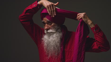 Low-Key-Studio-Lighting-Shot-Of-Senior-Sikh-Man-With-Beard-Tying-Fabric-For-Turban-And-Using-Salai-Needle-Against-Dark-Background