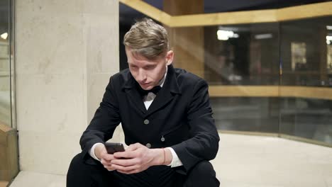 Young-businessman-in-black-suit-with-smartphone-sitting-on-stairs-in-hotel