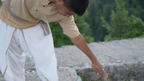 indian adult male doing yoga with closed eyes preparing for the pose touching the ground with hand and standing on barefoot on stone castle wall