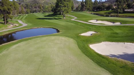 Vista-Aérea-De-4k-De-Un-Green-Con-Bunkers-De-Arena-Blanca-Y-Un-Estanque-En-Un-Campo-De-Golf-En-Los-ángeles,-California,-En-Un-Día-Cálido-Y-Soleado-Con-Montañas-Al-Fondo