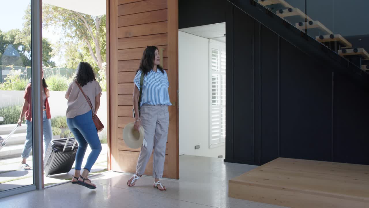 Diverse female friends entering home from trip, woman holding pivot door open and pulling suitcase