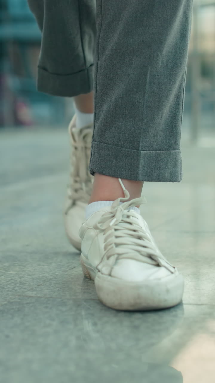 Leg view of individual in white sneakers and gray trousers dancing on polished tile floor with glass building and iron railing in background