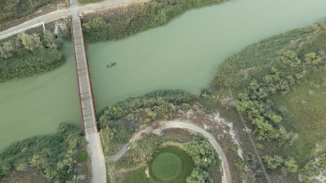 A-small-motorboat-floating-down-a-lazy-green-river---tilt-down-aerial-view