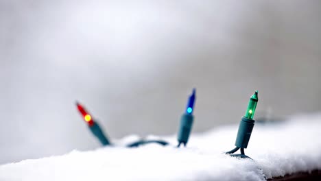 Close-up-of-blue,-green-and-orange-Christmas-lights-on-a-snowy-deck-with-a-blurred-background-and-snow-falling