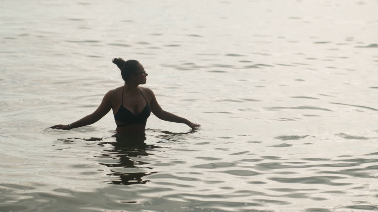 Kvinde i roligt hav til taljen, eftertænksom silhuet vader gennem blide krusninger i skumringen. Figur i sort badedragt praktiserer langsom vejrtrækning, poserer som en moderne danser og i meditation.