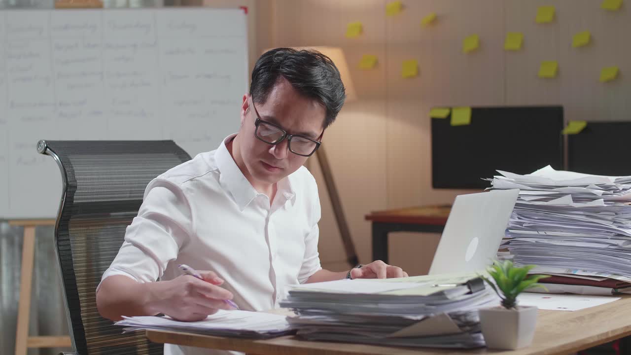 hombre asiático mirando la computadora portátil y tomando notas mientras trabaja con documentos en la oficina