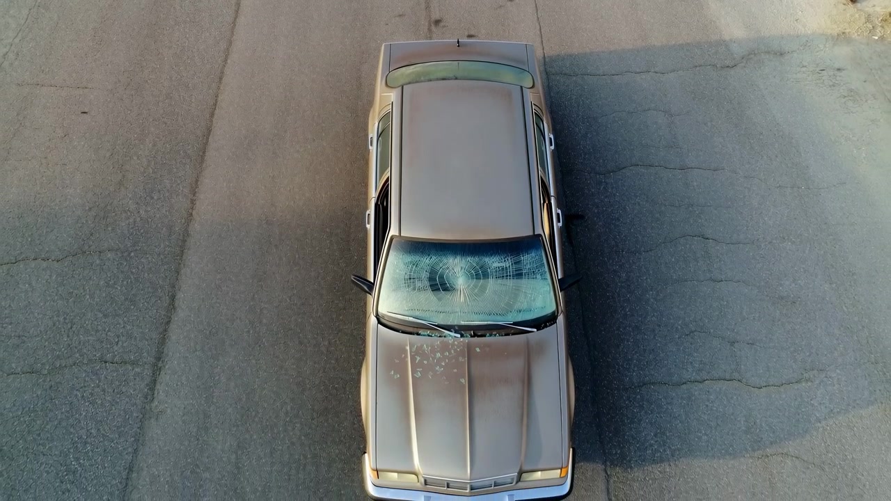 Vista cenital muestra un sedán estacionado con grietas de parabrisas tipo telaraña, fragmentos de vidrio roto en el capó, limpiaparabrisas desplazado, carrocería color tostado metálico y asfalto agrietado con manchas, juntas y tonos suaves y cálidos