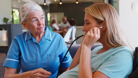 senior midwife visiting pregnant woman at home listening to heartbeat with stethoscope