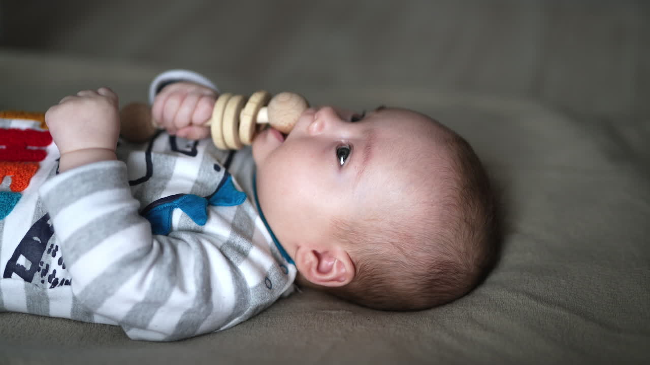 Little toddler took a wooden toy into mouth. Cute baby boy in striped costume lying on the bed. Side view. Grey backdrop. Close up.