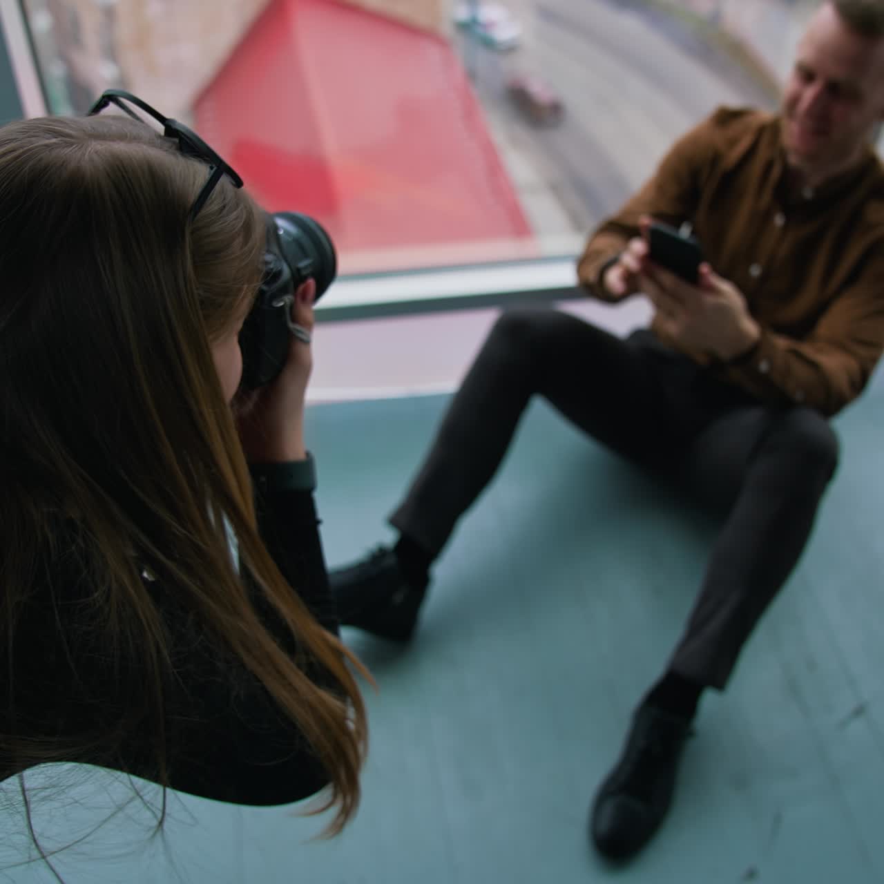 Photo session in office. Female photographer taking photos of businessman sitting on a floor with a mobile phone near the window with a city view