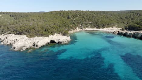 Vista-Aérea-De-Cala-Escorxada,-La-Playa-Virgen-En-Menorca,-España-Con-Agua-Azul-Y-Una-Colina-Cercana-Con-árboles-Verdes-Y-Cielo-Azul-Claro-En-El-Fondo