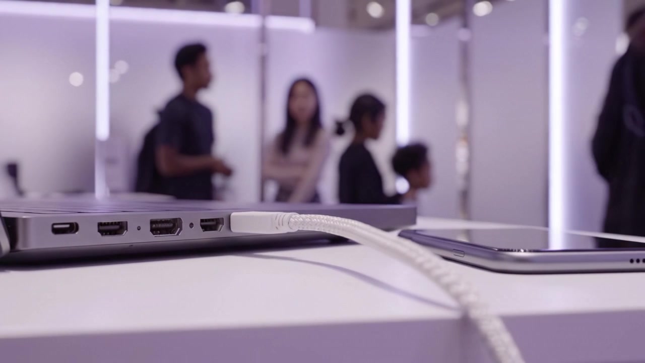 Close-up of Laptop Ports and Smartphone Charging Cables in a Modern Display Setting with People in the Background Engaged in Conversation