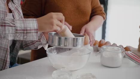 grandmother teaching granddaughter how to sift flour using kitchen strainer