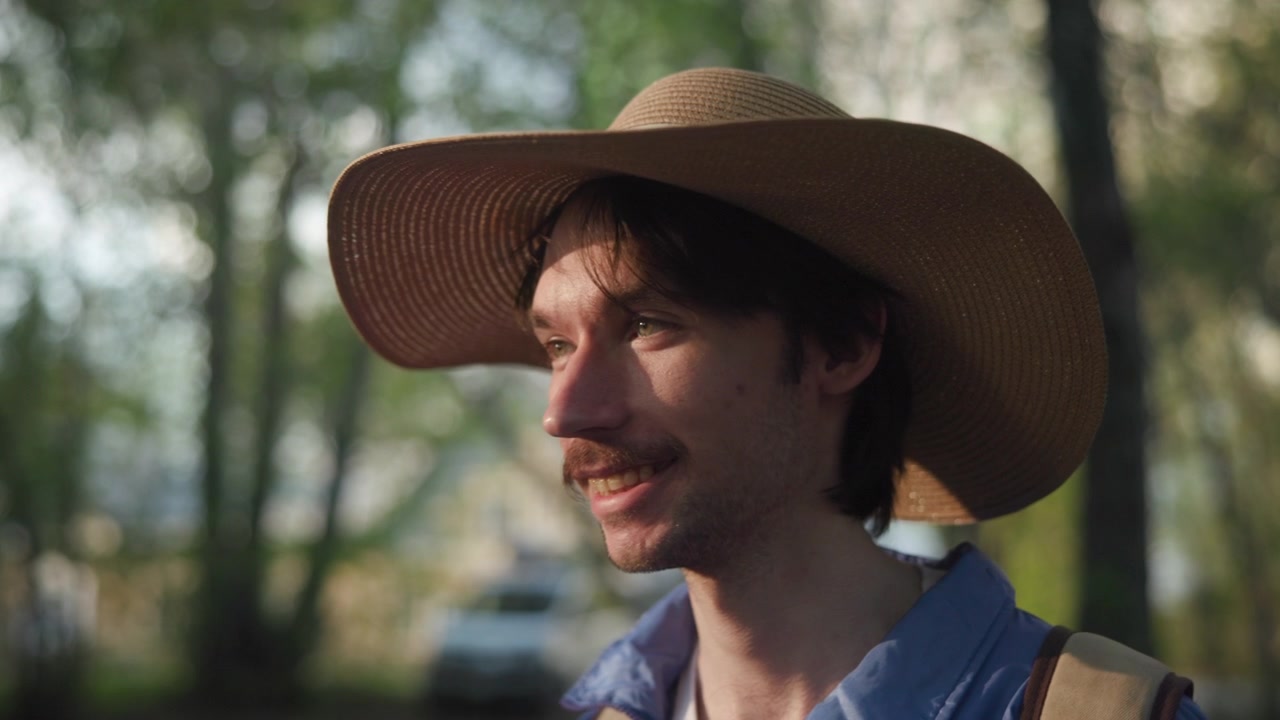Profilo laterale di un giovane uomo sorridente che indossa un ampio cappello di paglia e una camicia blu, in piedi in un parco o bosco illuminato dal sole con alberi verdi sfocati sullo sfondo.