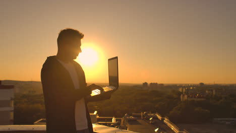 mit einem laptop auf dem dach mit city view und forex-chart. hacking und statistikkonzept. ein mann beim sonnenuntergang in slowmotion, der software-code auf einem laptops schreibt