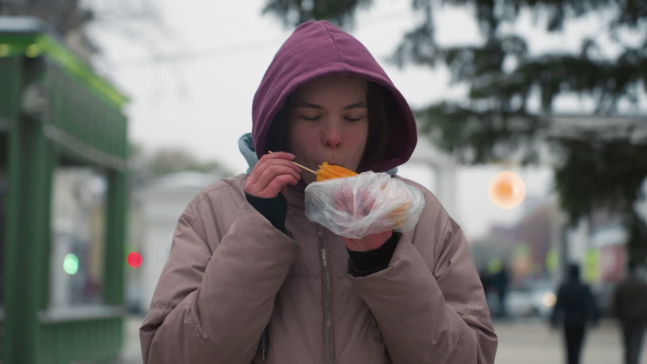 dama con chaqueta de invierno comiendo maíz en la calle al aire libre, sosteniendo maíz envuelto en plástico, disfrutando de comida callejera, clima frío, comida de invierno casual, chaqueta con capucha, entorno de parque urbano
