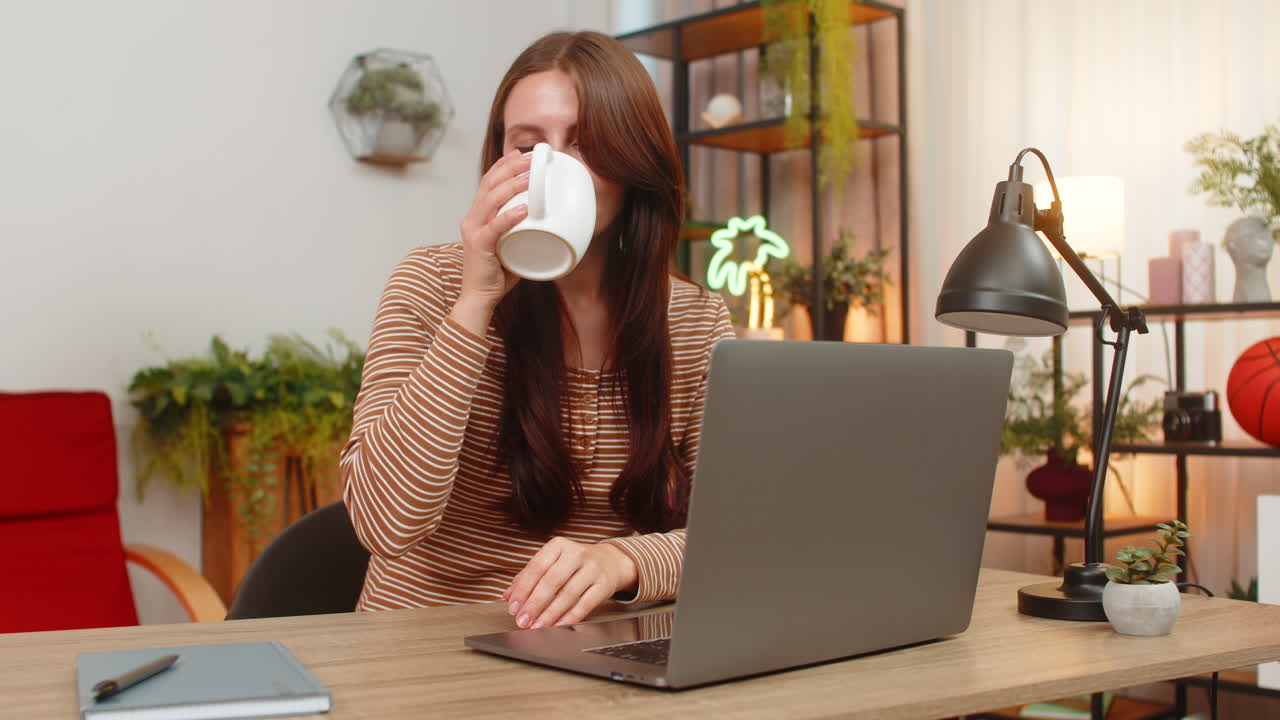 Una joven se calienta las manos con una taza de té caliente, mira el portátil y escribe un correo electrónico a un compañero con atención tranquila.
